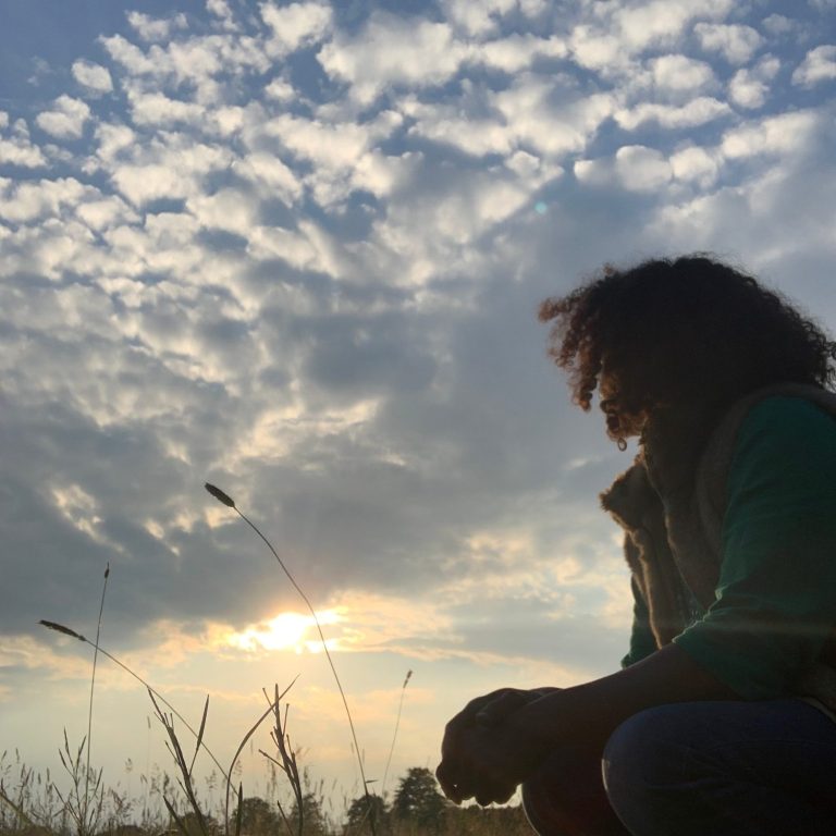 Eine Person mit lockigem Haar sitzt am Waldrand und betrachtet den Sonnenuntergang.